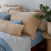 Linen Twin Fitted Sheets | a cluster of pillows surround deluxe sized shams in a pale honey color shown with blue sheets and bedding - a side table slightly visible in the shot against a white wall - no headboard.