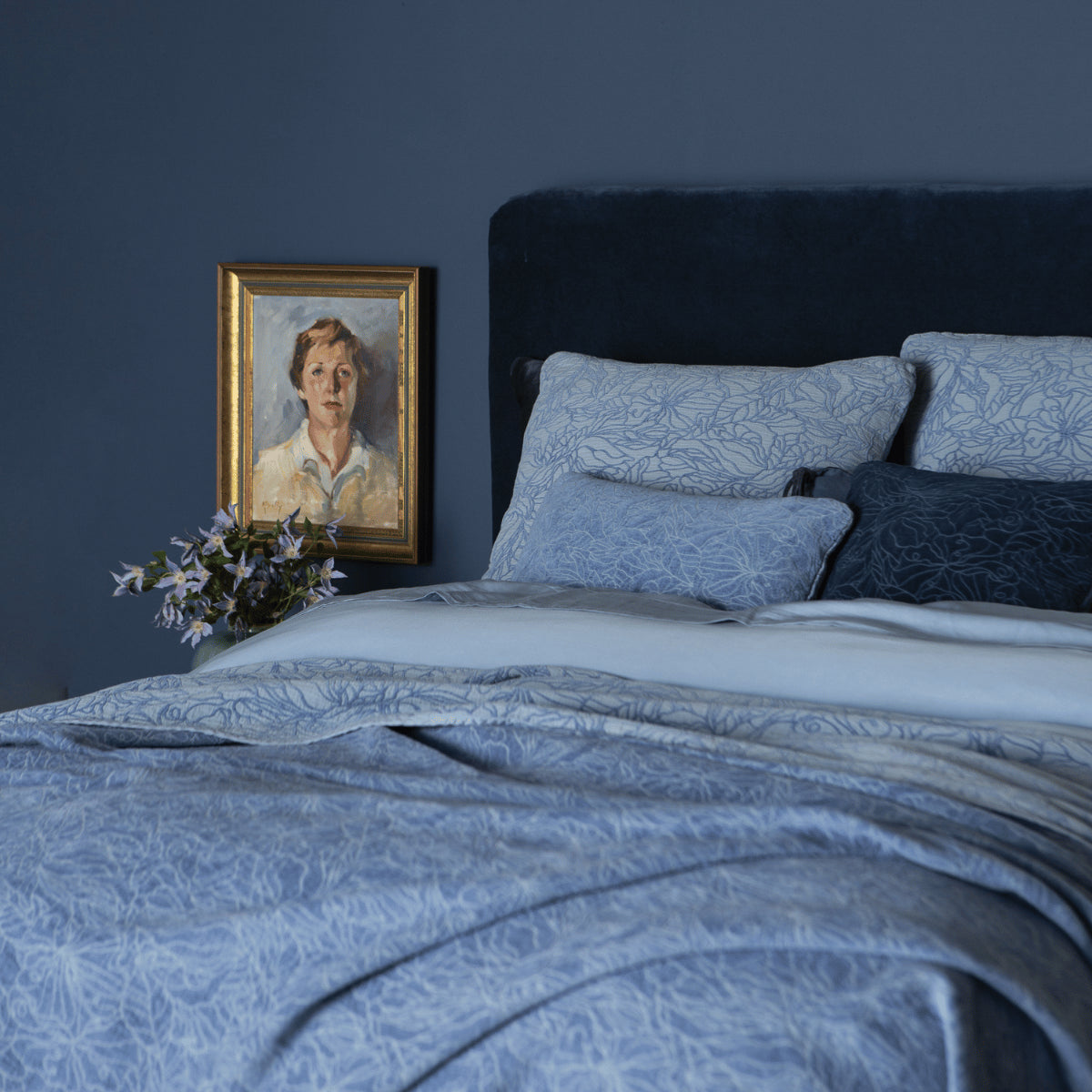 [allvariants]: a three-quarter view of a fully dressed bed in blue tones with a velvet headboard against a blue wall. 