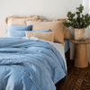 Linen Duvet Cover | Tahoe Blue | a bed dressed in all linen in blue and honey tones against a white wall with a side table holiding a small decorative bowl and vase of greenery.