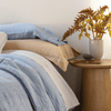 Linen Pillowcase (Single) | a blue pillowcase lays atop a honey-colored sham on a bed made in light blue linen bedding with a decorative side table between the bed and window.