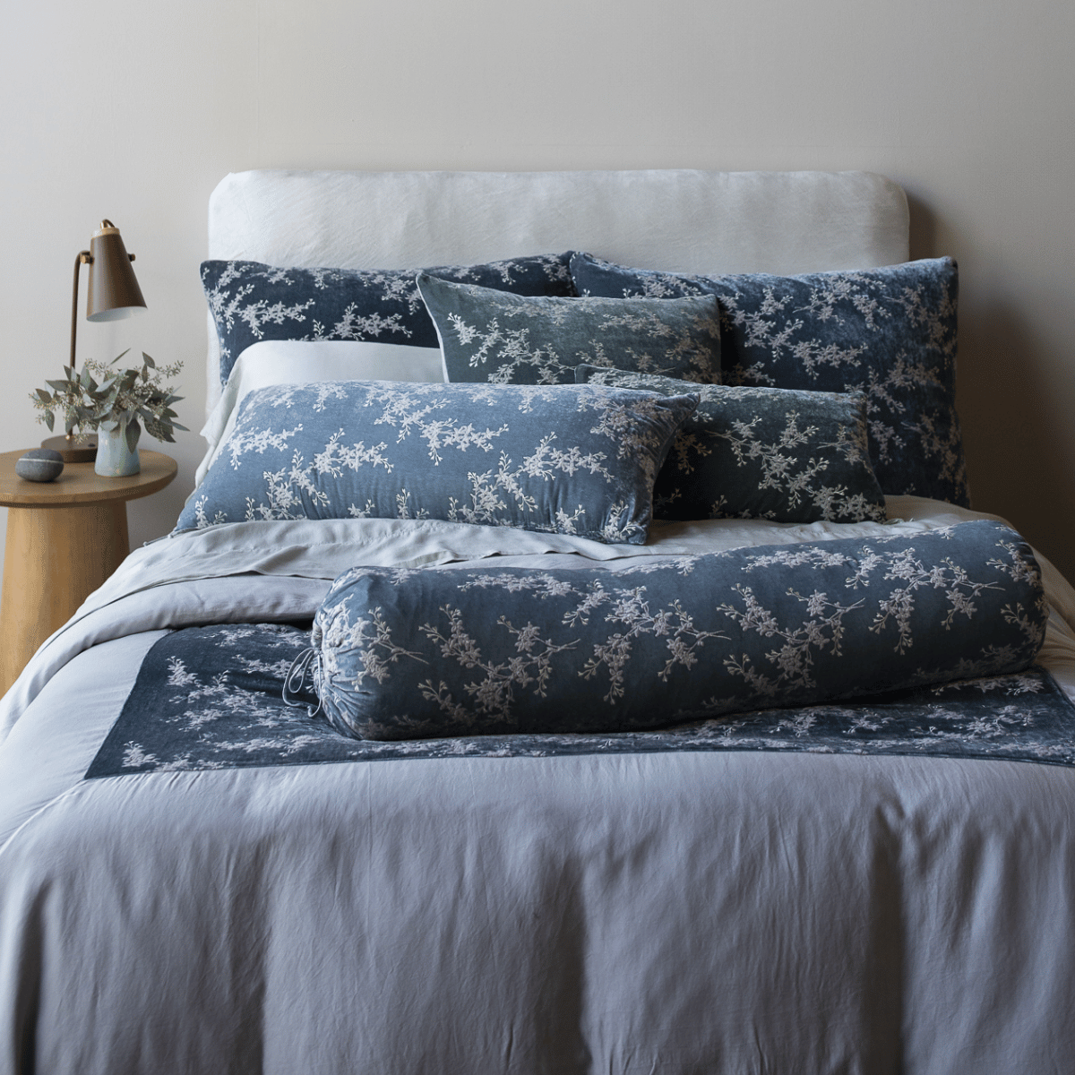 [allvariants]: a cluster of throw pillows and shams from the lynette and lynette luxe collections shown on a bed in light blue and grey tones against a neutral toned wall.