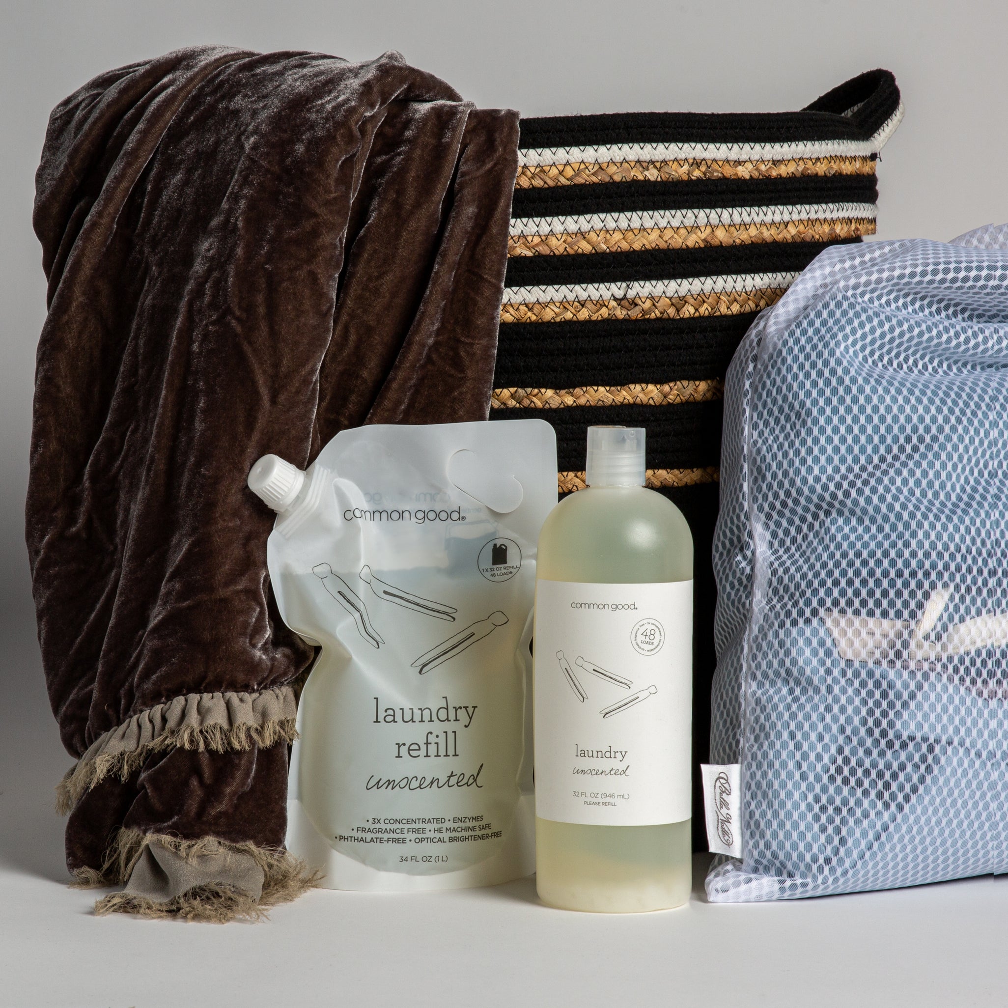 A shot of laundry products including a detergent refill, detergent bottle and mesh laundry bag shown with a basket and silk velvet blanket. 