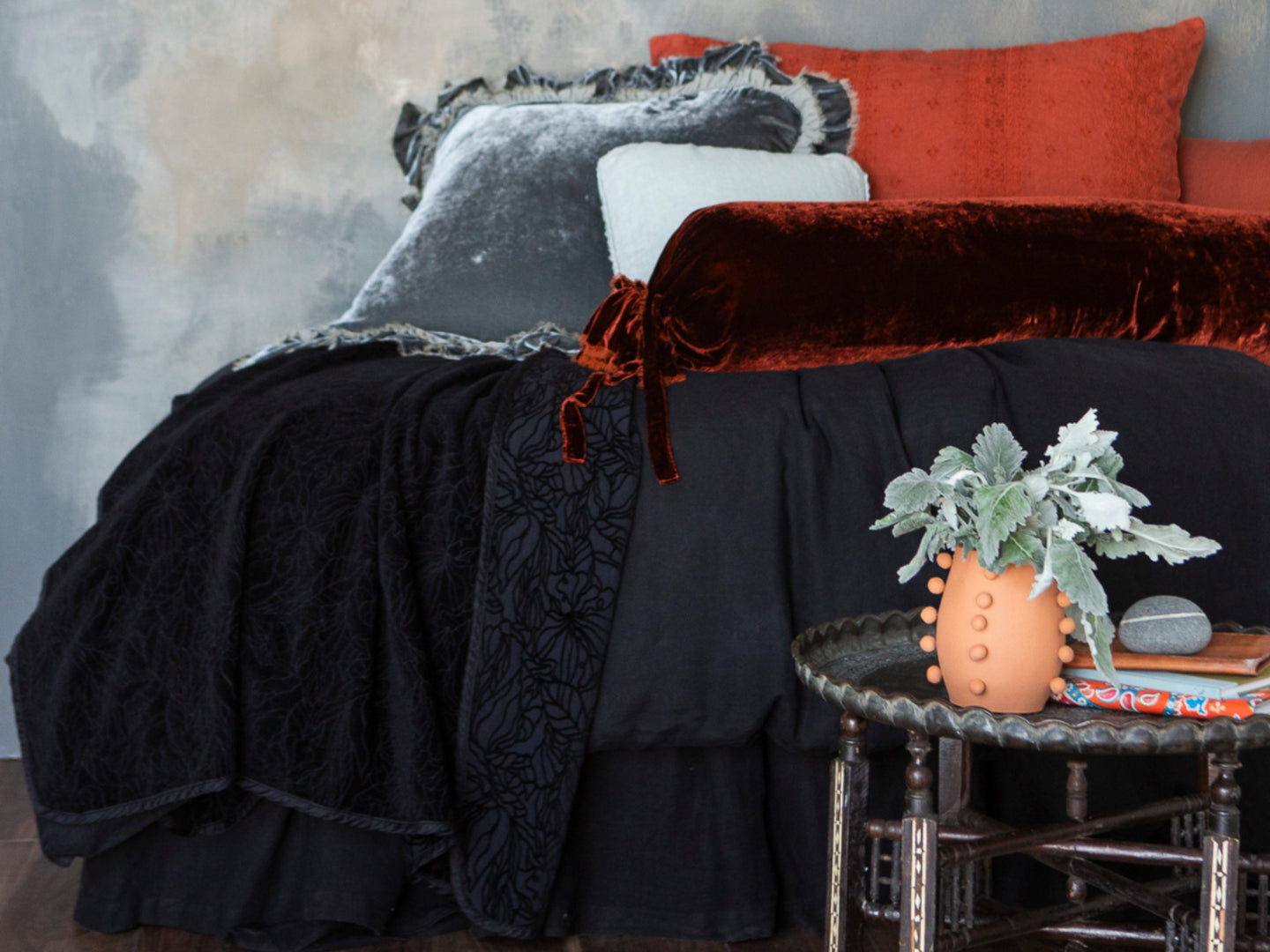 A dark bed dressed in black, grey and rust tones, a side table with books and greenery accentuates the bedding.