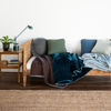 Carmen Blanket | A daybed bench against a white wall and dark wood flooring. A neutral rug ties in with the bench and side table. An assortment of throw pillows and blanket in grey and blue tones is the focus.