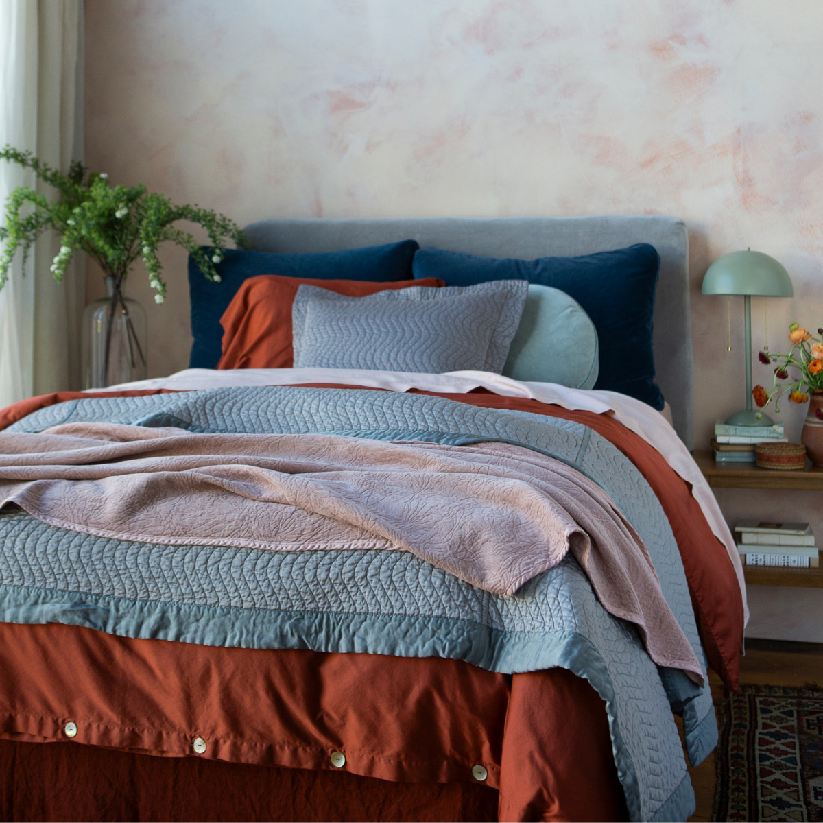 A colorful bed shown from a slight angle against a warm toned limewash wall with greenery on each side of the headboard. 