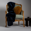 Silk Velvet Quilted Blanket | Midnight | a dark blue quilted silk velvet throw blanket with a round pillow in a golden tone on an ornate chair with a small side table.