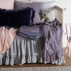 Linen Whisper Crib Skirt | French Lavender | a close-up of a crib skirt and baby comforter from the linen whisper collection on a crib with several pillows and other baby blankets.