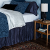 Paloma Bed Skirt | dark blue bed skirt on a bed dressed in other blue tones and dark floor and rug with coordinating side table.