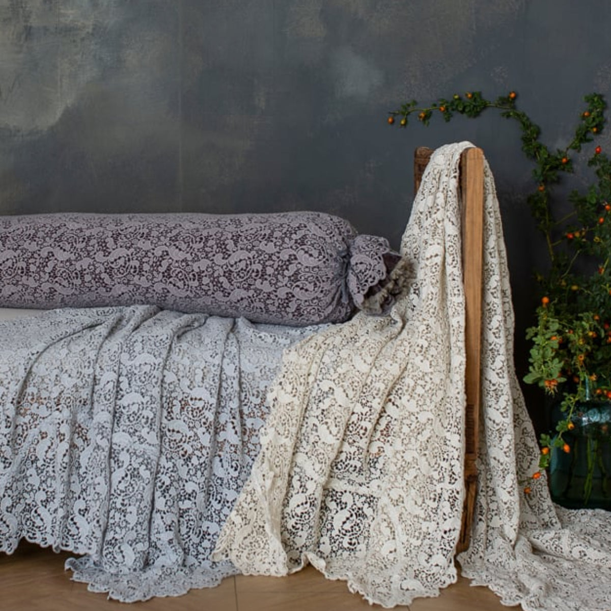 [allvariants]: lace bolster cover on a bench with lace bed scarves against a limewash background. 