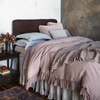 Linen Whisper Duvet Cover | 3/4 view of a bed dressed in mostly linen pieces in lavender and warm grey tones against a dark limewash wall with moody lighting.