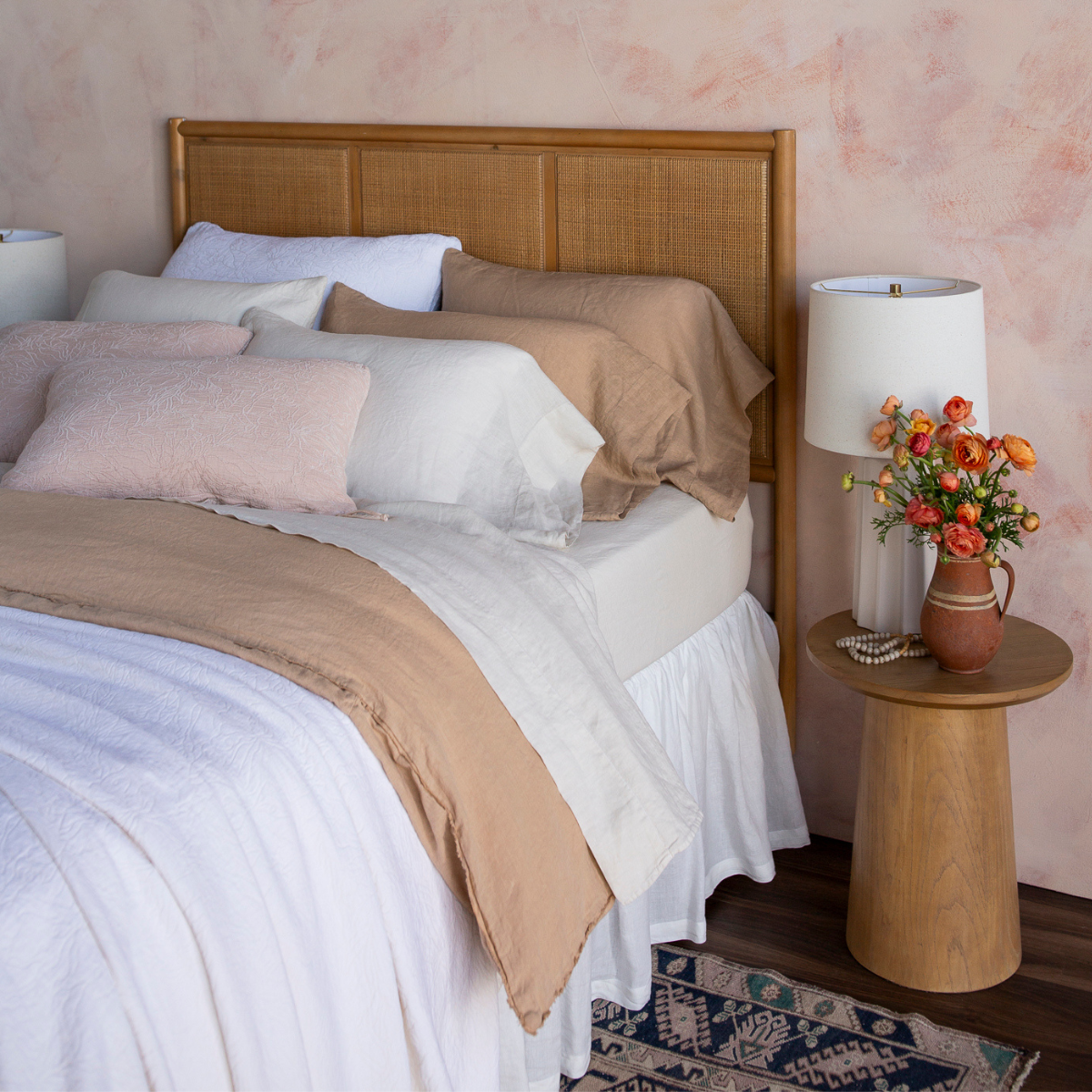 [allvariants]: a three-quarter view of a fully dressed bed in light tones with a rattan headboard against a pink-toned wall. 