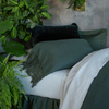 Linen Pillowcase (Single) | Juniper | a lace-tirmmed linen pillowcase in juniper, a dark green, on a bed with other green and light-colored tones surrounded by lots of green plants.