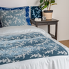 Lynette Luxe Duvet Cover | Cloud | shown from the foot of the bed, an embroidered silk velvet panel inset into a tencel duvet cover and matching shams are shown with a side table holding a notepad and pen, glass of water and potted plant.