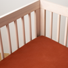 Autumn | A close-up corner view of a Madera Luxe Crib Sheet in Autumn, a rich terracotta tone, fitted on a crib.