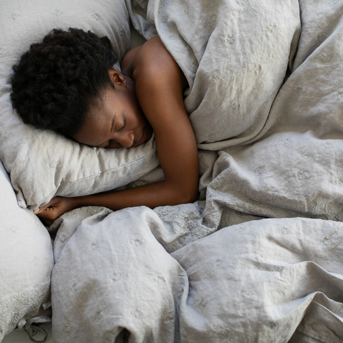 A woman sleeping on an embroidered midweight dressed bed in a light grey tone. 