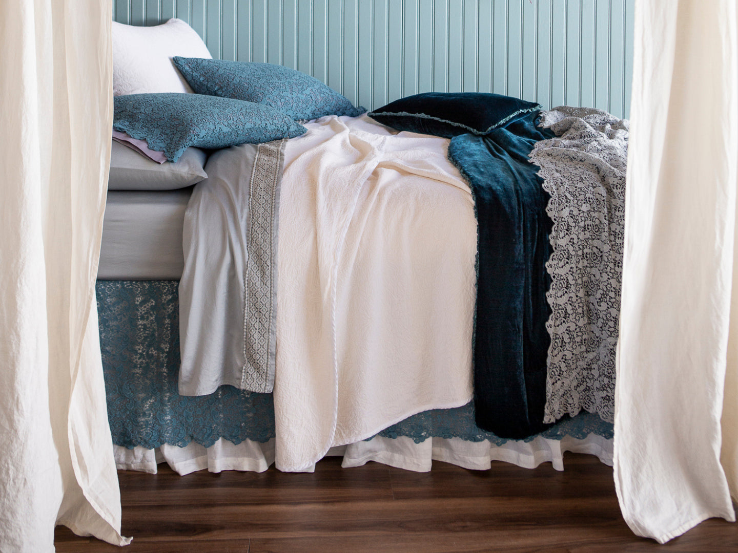 a bed dressed in layers of lace and jewel tones, shown from the side with curtains in the foreground and a light blue panelled wall in the back. 